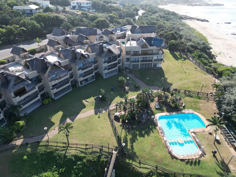 Aerial view of a coastal apartment complex featuring thatched roof architecture, expansive landscaped gardens, and a communal swimming pool area near the beach.