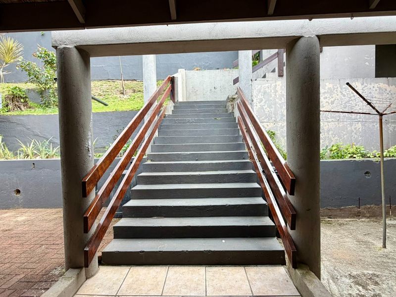 An outdoor concrete staircase with timber handrails leading up from a paved patio area, flanked by concrete columns and retaining walls.