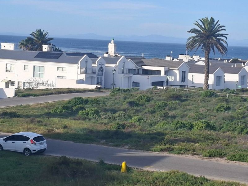 An elevated aerial view of a coastal residential area featuring white buildings, paved roads, and surrounding natural vegetation with ocean views.