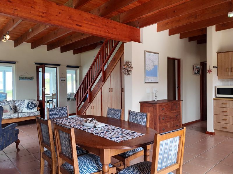 Open-plan living and dining area featuring exposed timber ceiling beams, tiled flooring, a timber staircase, and a functional kitchen with built-in appliances.