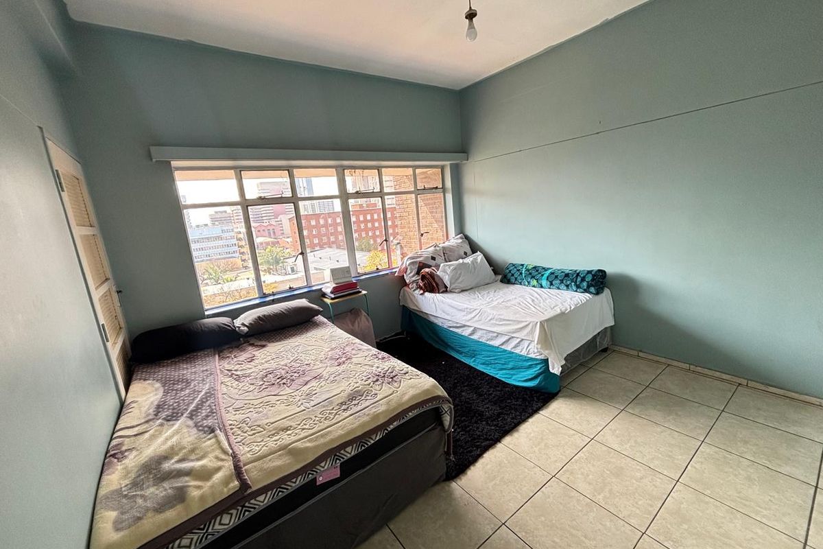 A spacious bedroom featuring large steel-framed windows with city views, ceramic tiled flooring, and built-in storage space.