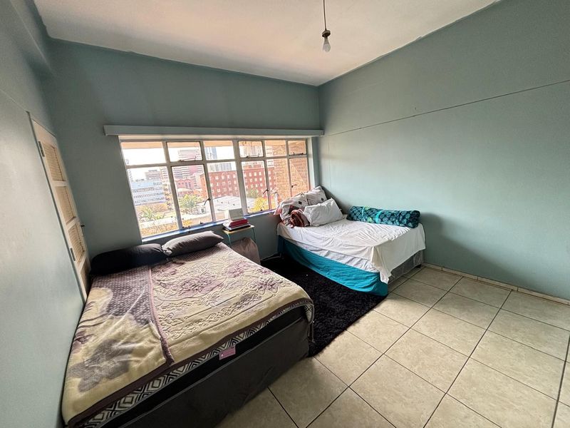 A spacious bedroom featuring large steel-framed windows with city views, ceramic tiled flooring, and built-in storage space.