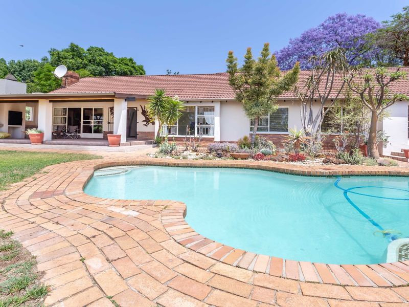 A well-maintained rear garden featuring a swimming pool with brick coping, a covered patio area, and established landscaping against the house facade.