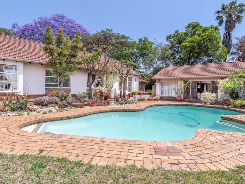 A well-maintained outdoor pool area featuring brick paving, established garden landscaping, and a clear view of the rear house elevation.