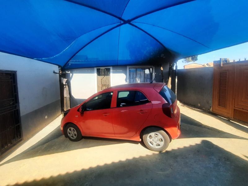 A paved driveway area featuring a large blue shade sail for vehicle protection, enclosed by perimeter walls and a secure gate.