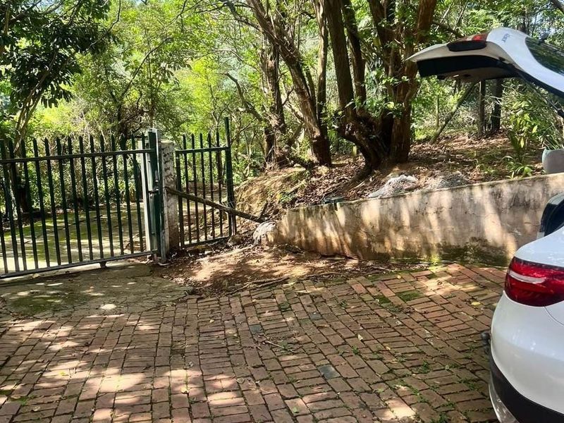 A view of the property entrance featuring brick paving, a metal security gate, and a retaining wall surrounded by mature trees.
