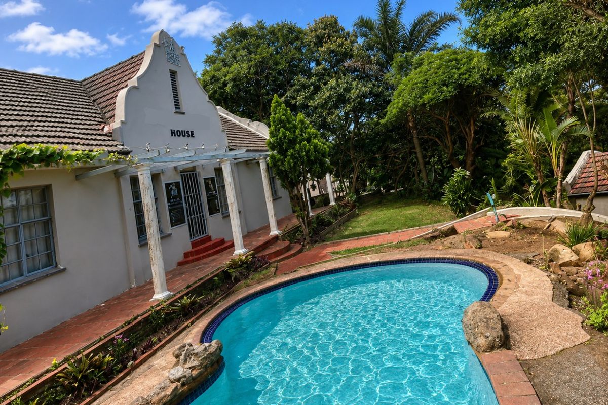 A view of the swimming pool area featuring a gabled house facade, a covered patio with columns, and established garden surroundings.
