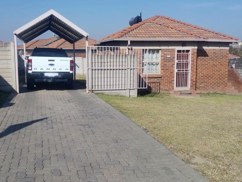 Front view of a single-story brick house featuring a paved driveway, covered carport, security gate, and a well-maintained front lawn.