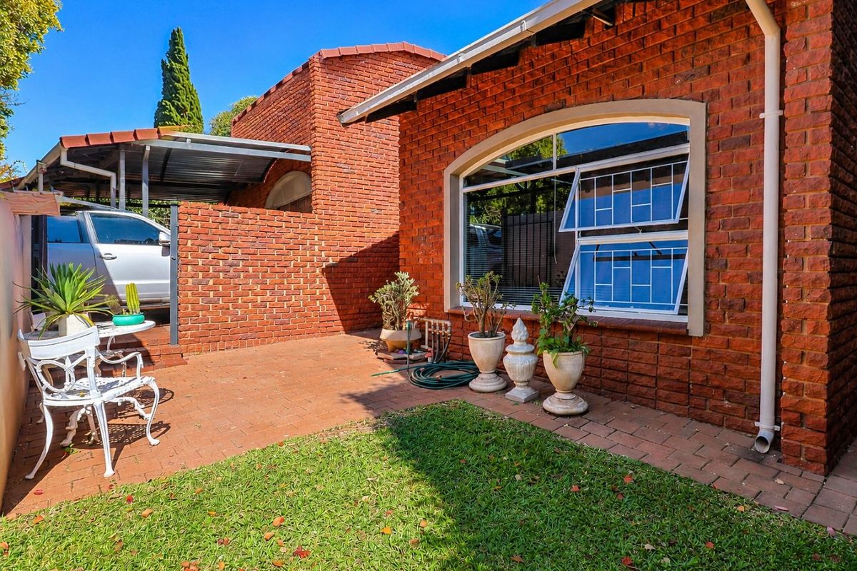 A private rear courtyard featuring a brick facade, paved patio area, and a small lawn, adjacent to a covered carport.