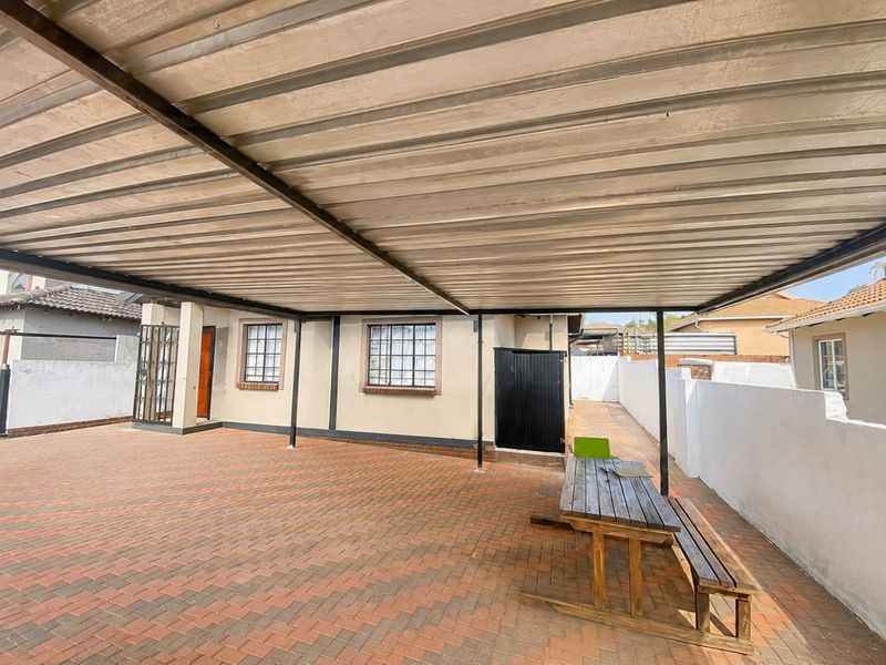 A covered outdoor patio area featuring brick paving, a corrugated metal roof structure, and secure window bars, providing a functional space for outdoor living.