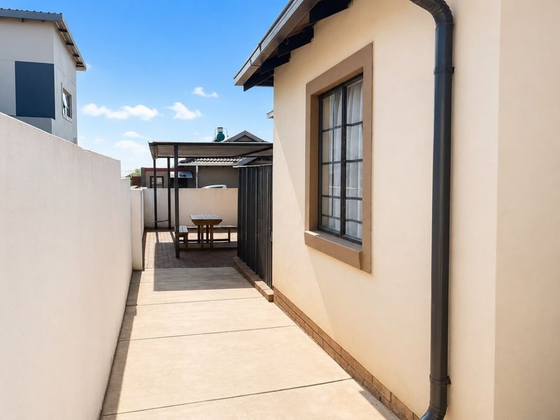 A side walkway leading to a paved rear patio area with a covered structure, featuring clean exterior finishes and boundary walling.