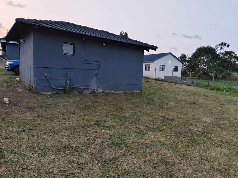 Rear view of a dark-painted residential structure with exposed exterior plumbing and a tiled roof, set on a large grassed plot.
