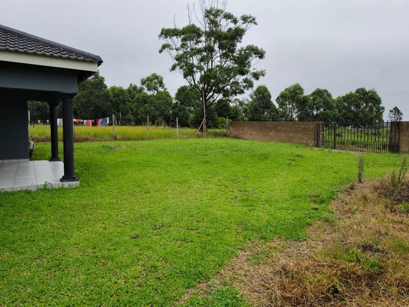 A spacious backyard featuring a covered tiled patio with concrete columns, overlooking a large grass lawn enclosed by a perimeter wall and gate.