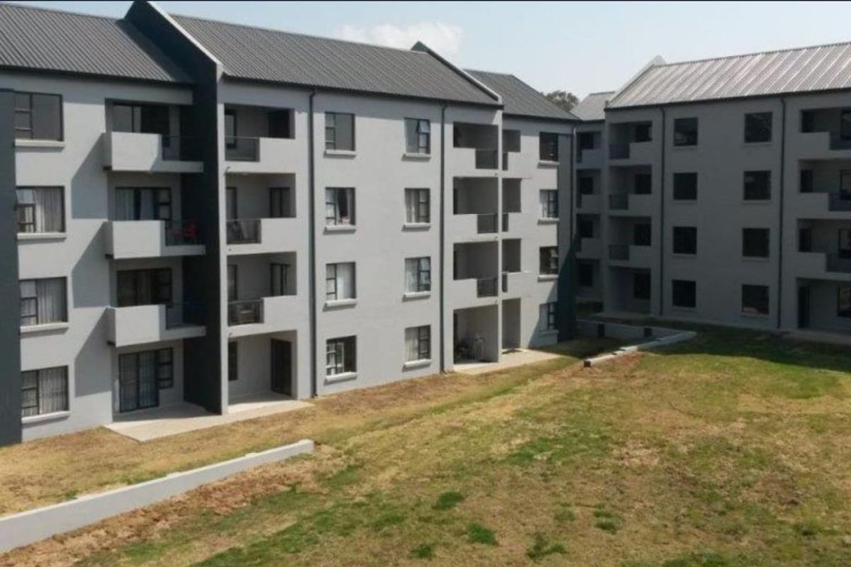 Exterior view of a modern multi-story apartment complex featuring grey facades, metal roofing, and private balconies overlooking a communal lawn area.