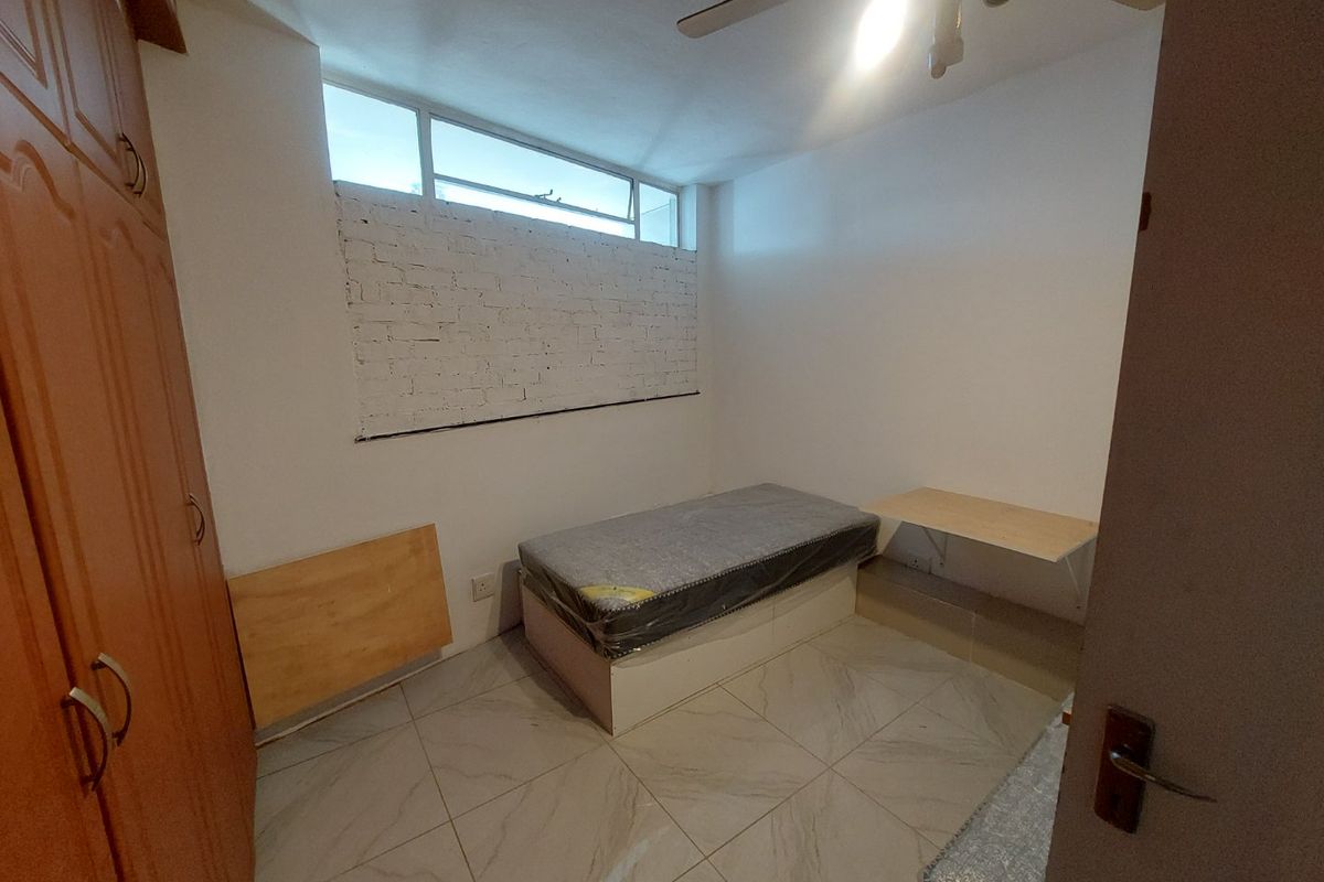 A compact bedroom featuring tiled flooring, a built-in wardrobe, a ceiling fan, and a high-level window with an exposed brick feature wall.