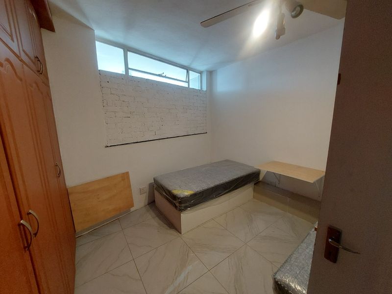 A compact bedroom featuring tiled flooring, a built-in wardrobe, a ceiling fan, and a high-level window with an exposed brick feature wall.