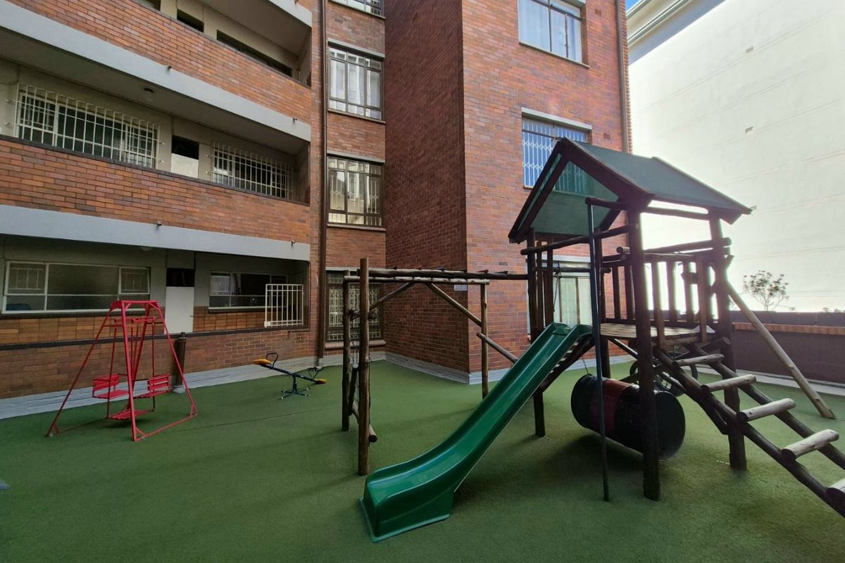 A communal outdoor area featuring a brick apartment building facade with security bars and an artificial turf surface with playground equipment.