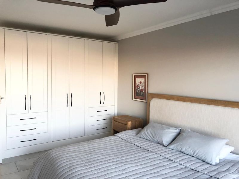 A well-maintained bedroom featuring extensive white built-in cupboards, a modern ceiling fan, and neutral tiled flooring.