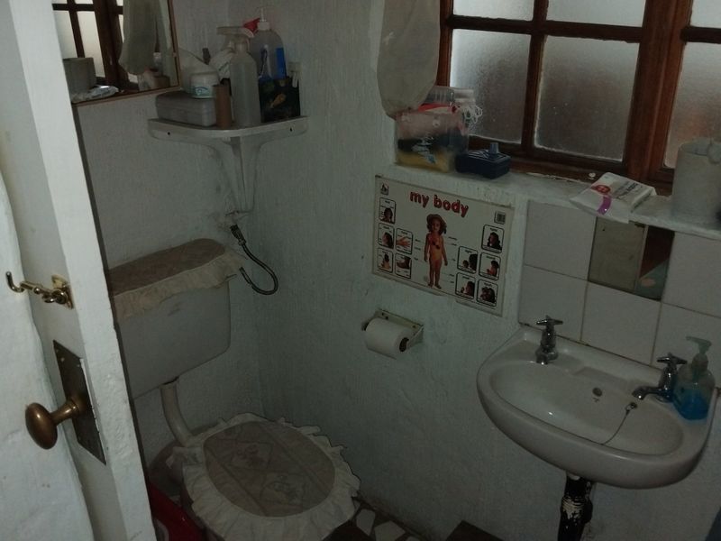 A compact bathroom featuring a wall-mounted basin and toilet, with dated finishes and a timber-framed window.