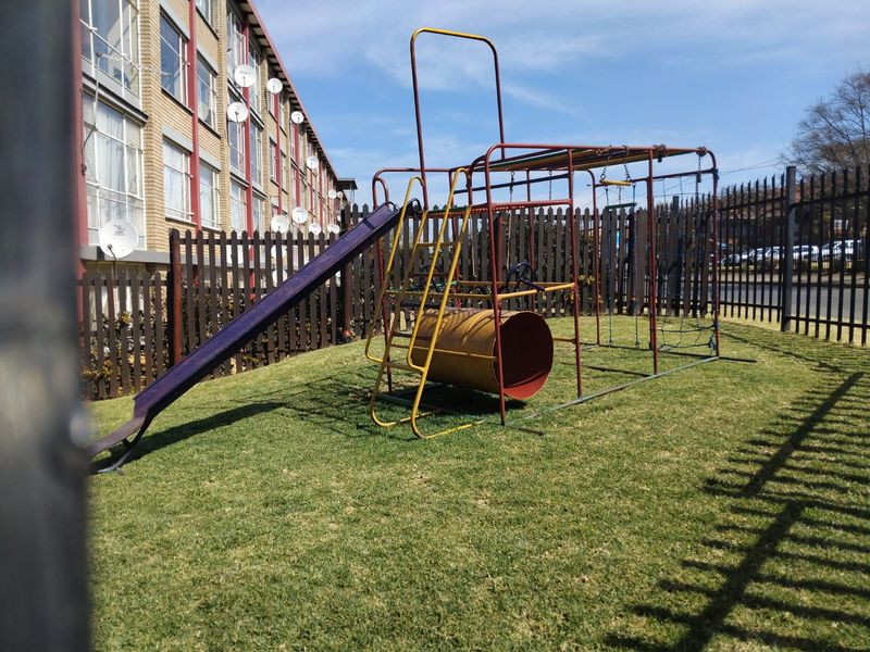 Communal garden area featuring a lawn and fixed playground equipment, enclosed by metal perimeter fencing adjacent to the apartment building.