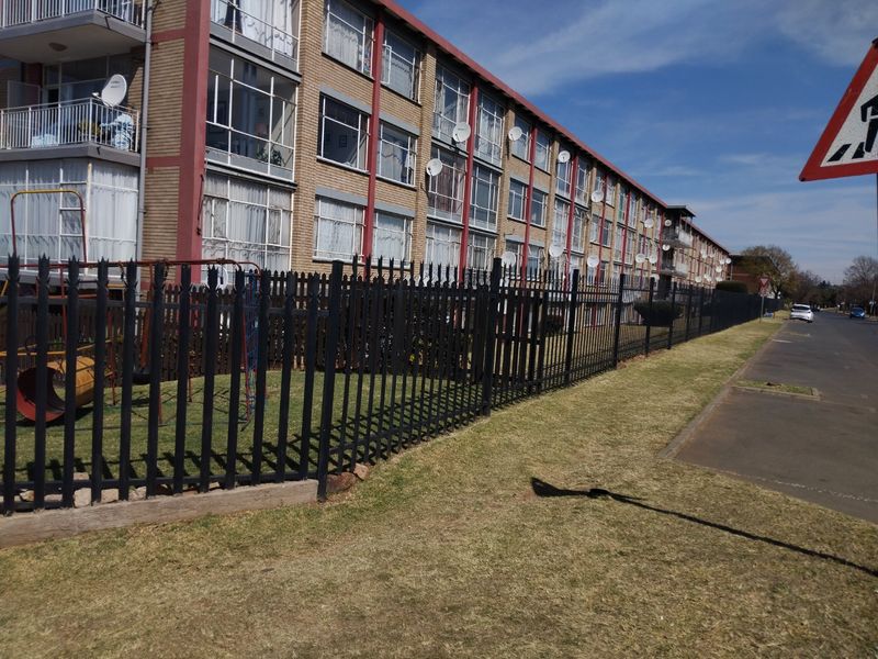 Exterior view of a multi-story apartment building featuring brick facade, balconies, and secure metal perimeter fencing along the street front.