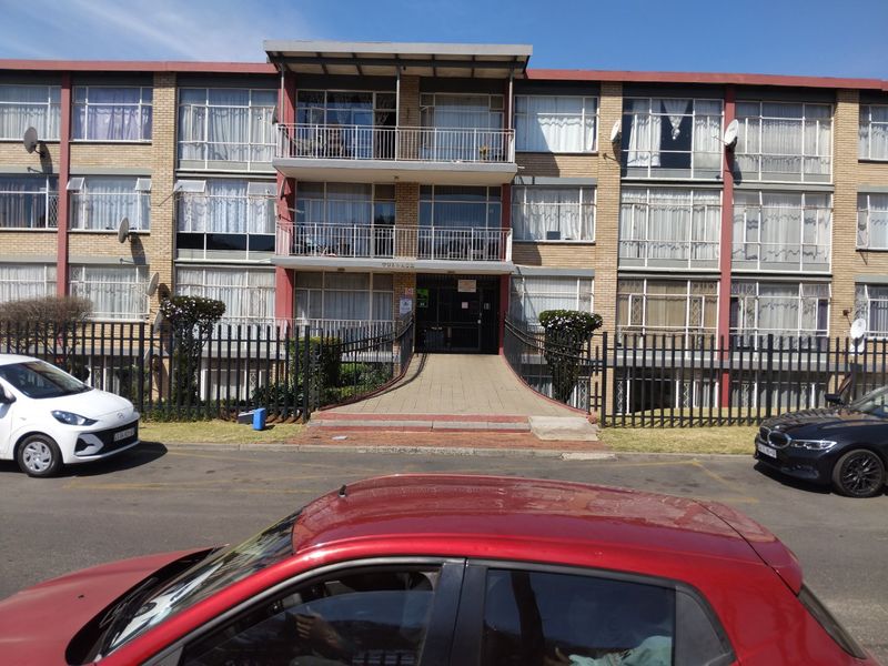 Front elevation of a multi-story apartment building featuring brick facade, metal balcony railings, and a paved entrance walkway with security fencing.