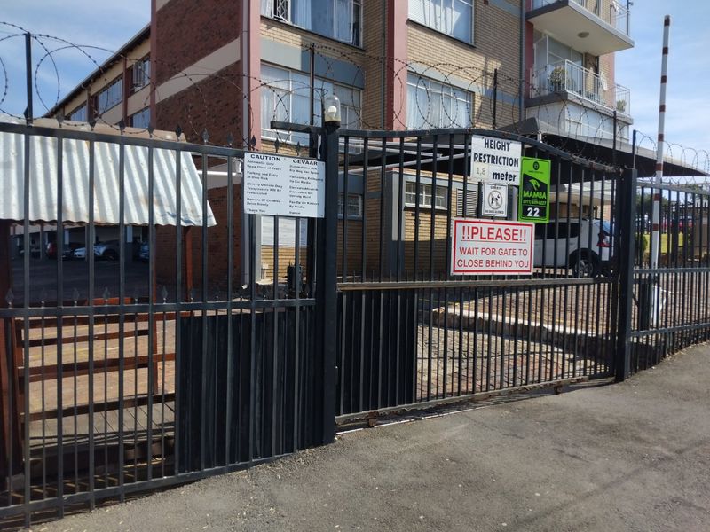 Front entrance of the apartment complex featuring a secure automated gate, razor wire fencing, and visible parking area.