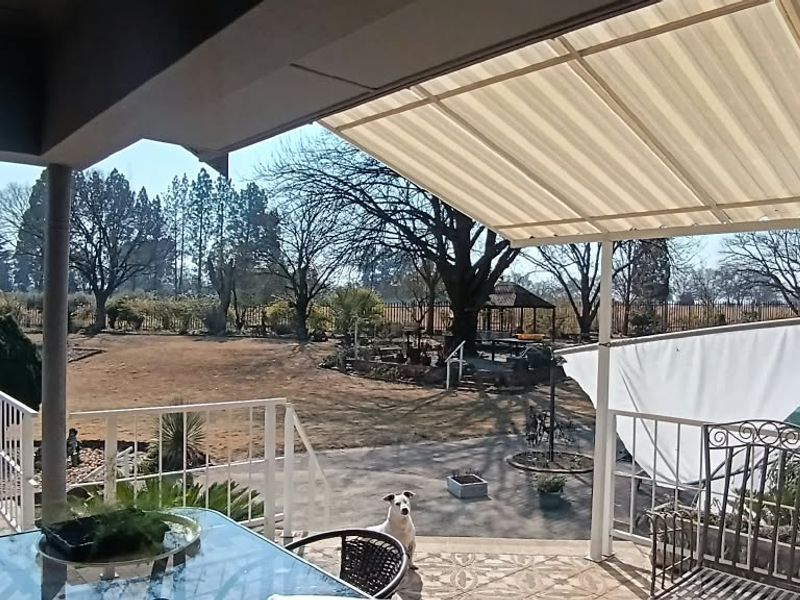 A covered outdoor patio area featuring tiled flooring and metal railings, overlooking a spacious garden with mature trees.