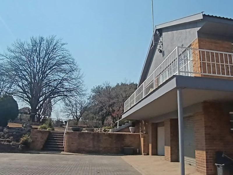 Rear view of a multi-level brick house featuring a spacious balcony, paved driveway, and tiered garden landscaping with stone retaining walls.