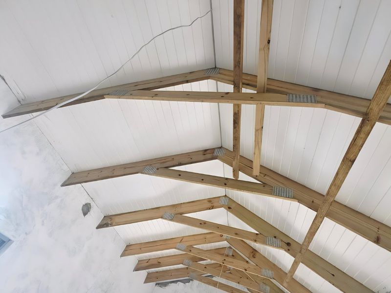 An unfinished interior space featuring exposed timber roof trusses, vaulted tongue and groove ceilings, and large aluminium window and door frames.