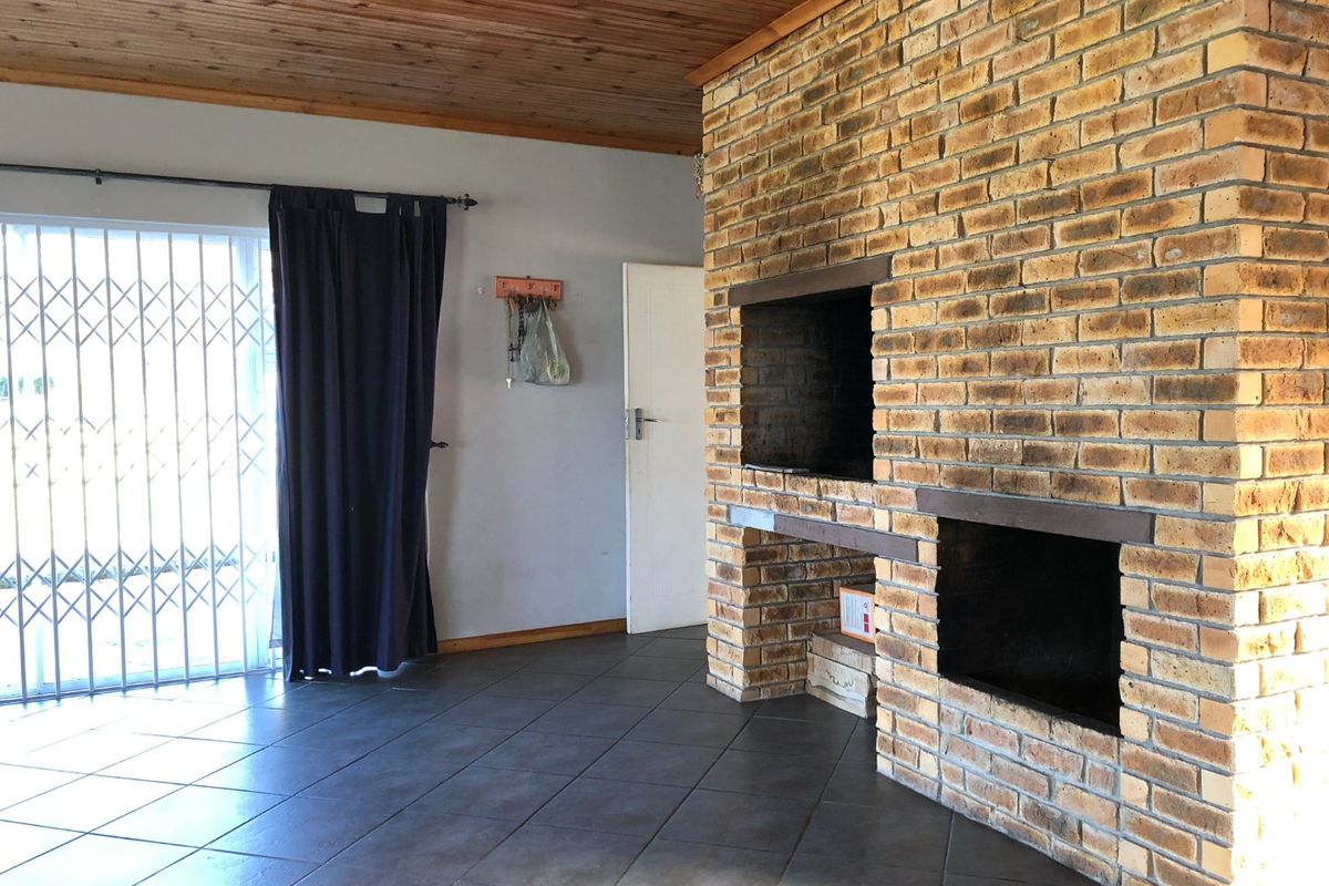 Spacious living area featuring a prominent brick fireplace with built-in braai, tiled flooring, and a timber-clad ceiling, offering a rustic farm-style aesthetic.