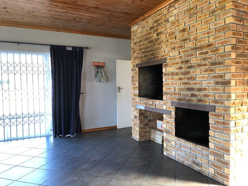 Spacious living area featuring a prominent brick fireplace with built-in braai, tiled flooring, and a timber-clad ceiling, offering a rustic farm-style aesthetic.