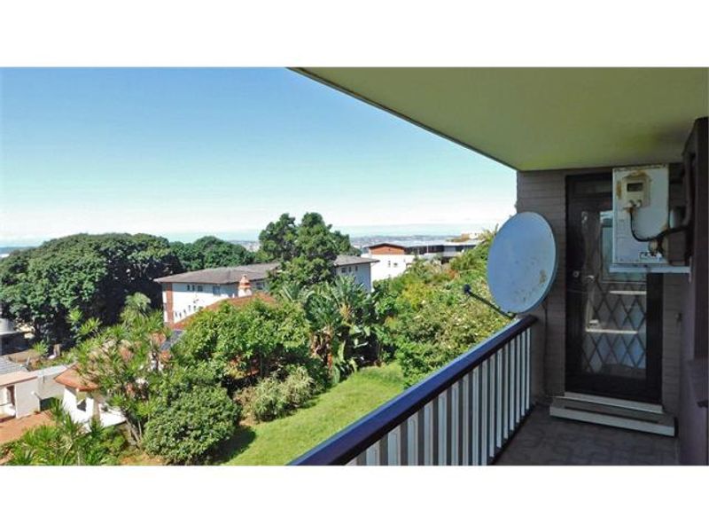 A covered balcony area featuring tiled flooring, a safety railing, and an external door, offering an elevated view of the surrounding neighborhood and greenery.