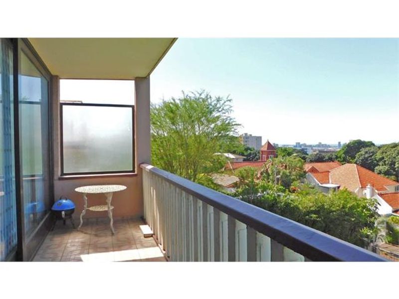 A covered balcony featuring tiled flooring and a railing, offering an elevated view of the surrounding neighborhood and greenery.