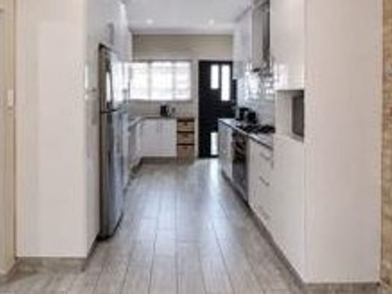 A modern galley-style kitchen featuring white cabinetry, tiled flooring, and integrated appliances, leading to an exterior door.