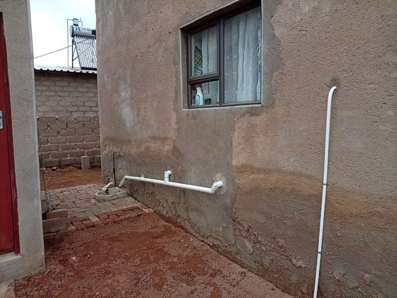 Rear exterior view showing unfinished plaster walls, external plumbing, a solar water heater, and a small concrete basin area.