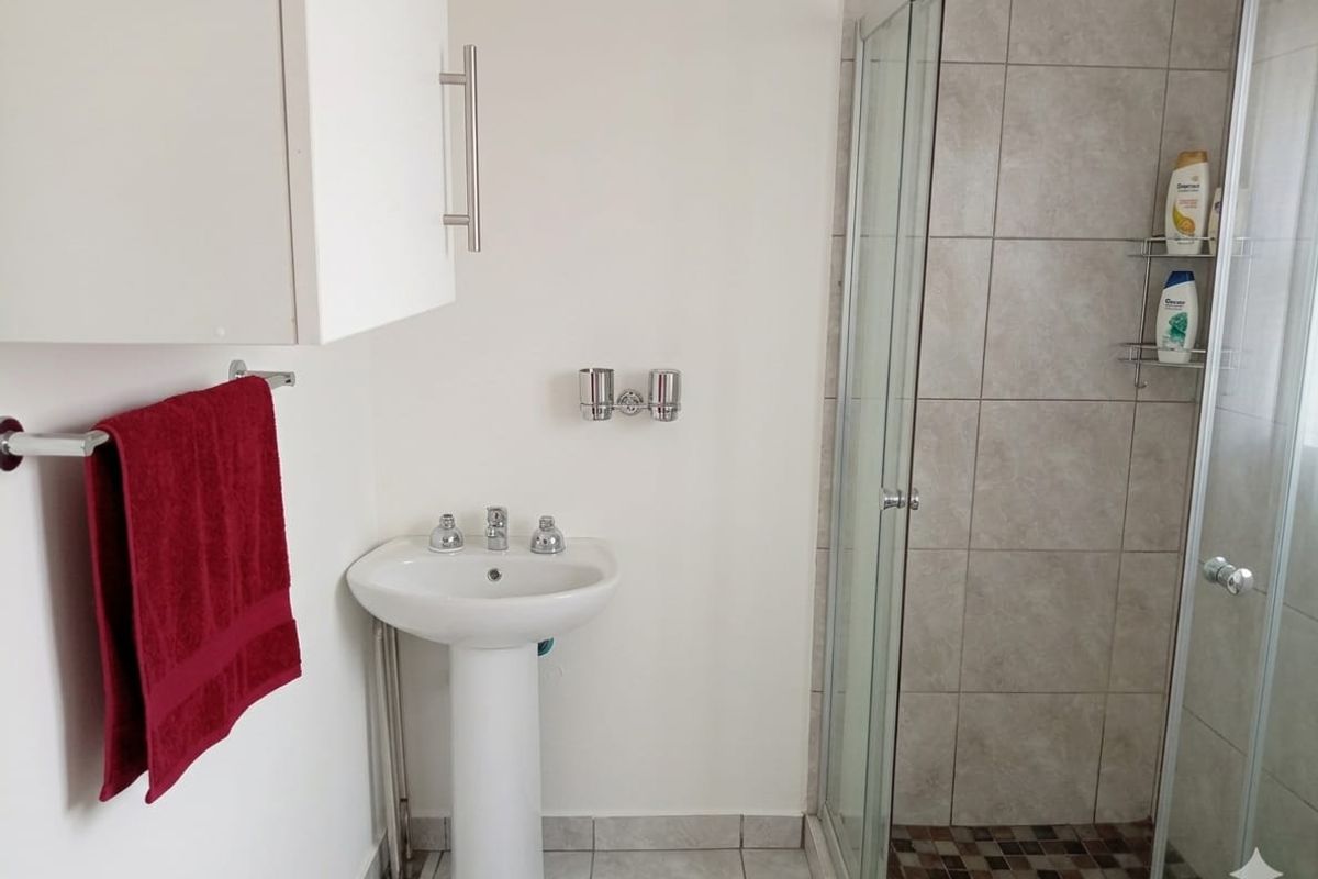 A functional bathroom featuring a pedestal basin, a glass-enclosed shower with tiled walls, and neutral floor tiling.