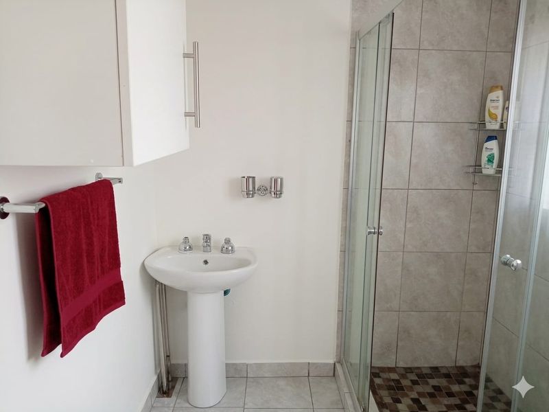 A functional bathroom featuring a pedestal basin, a glass-enclosed shower with tiled walls, and neutral floor tiling.