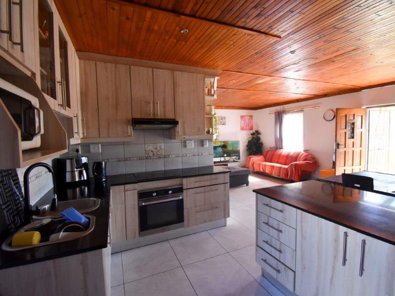 Open-plan kitchen and living area featuring light wood cabinetry, dark stone countertops, integrated appliances, and a distinctive timber-paneled ceiling.