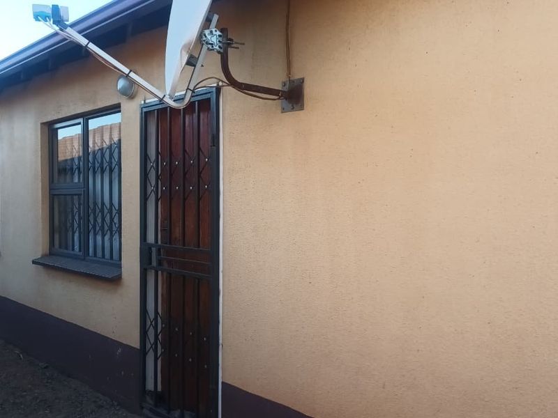 Side view of the property exterior featuring a secure entrance door with a metal gate, window security bars, and a mounted satellite dish.