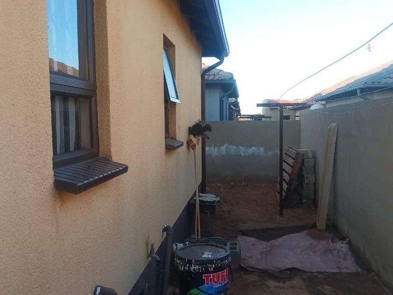 A side exterior view of the property showing plastered walls, aluminium window frames, and a boundary wall with an unfinished ground area.