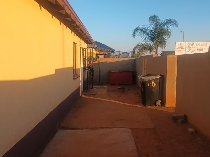A view of the rear exterior area of the house featuring a perimeter wall, security gate, and a mix of paved and unpaved ground.