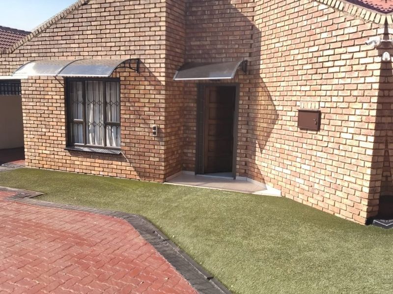 Front elevation of a face brick house featuring a paved driveway, artificial lawn, and protective window awnings over the entrance and window.