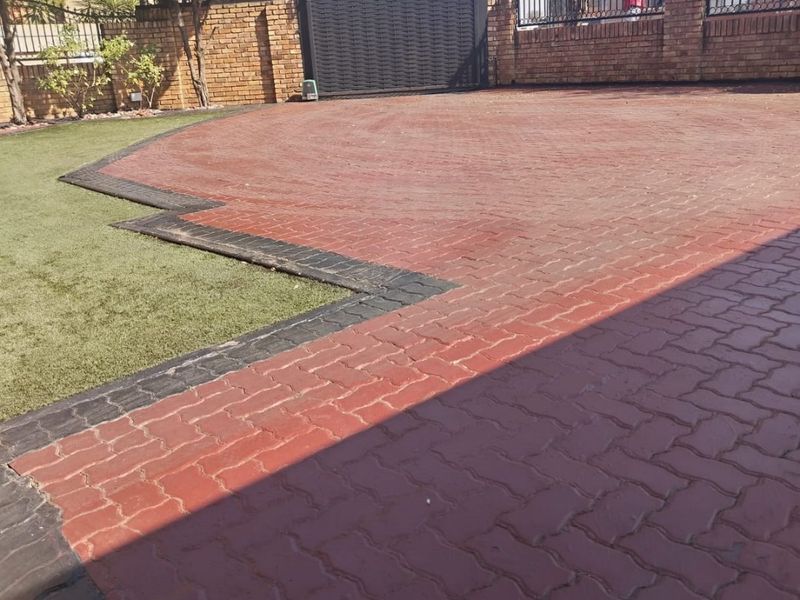 A well-maintained front driveway featuring red and black interlocking pavers, artificial grass, and a secure metal gate with brick perimeter walling.
