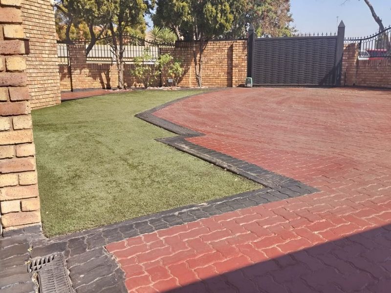 A well-maintained front exterior featuring a paved driveway, artificial lawn, brick boundary walls, and a secure metal gate.