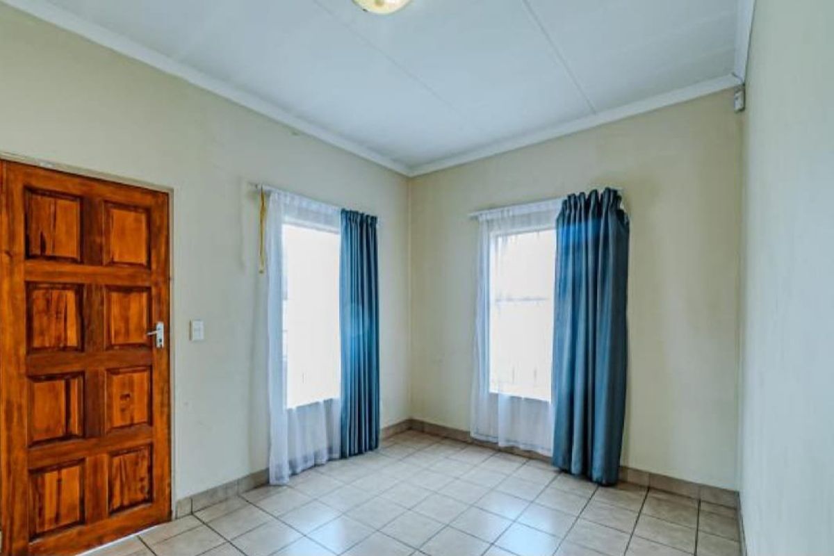 A bright, empty bedroom featuring tiled flooring, a solid wooden door, and dual windows with curtains.