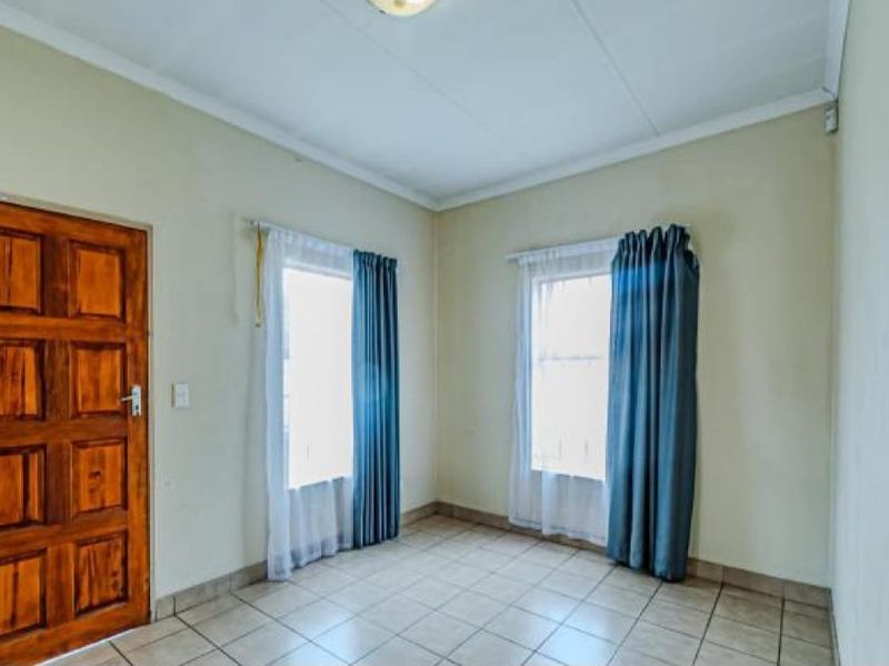 A bright, empty bedroom featuring tiled flooring, a solid wooden door, and dual windows with curtains.