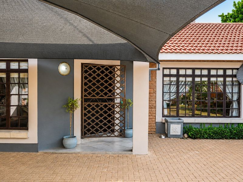Front entrance area featuring a paved driveway, shade sail covering, security gate, and well-maintained brick and rendered facade.