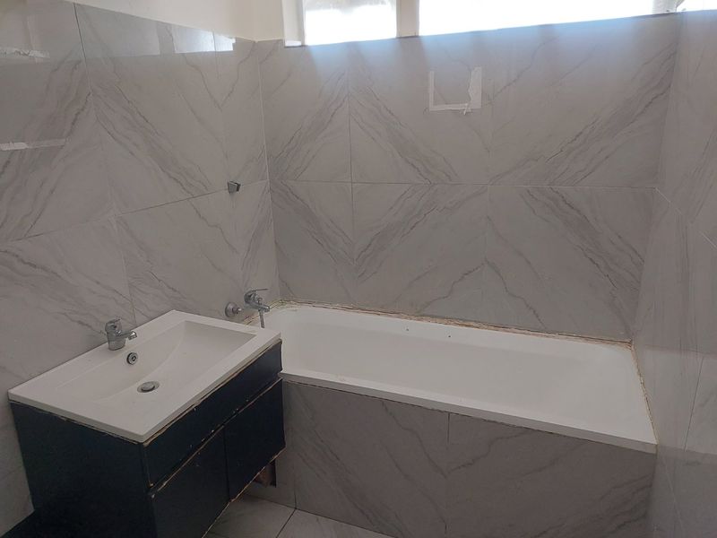 A bathroom featuring a built-in bathtub and vanity basin with grey tiled walls. The space requires minor repairs to finishes and sealant.