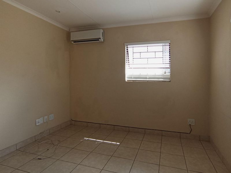 An empty bedroom featuring tiled flooring, a wall-mounted air conditioning unit, and windows fitted with security bars and venetian blinds.